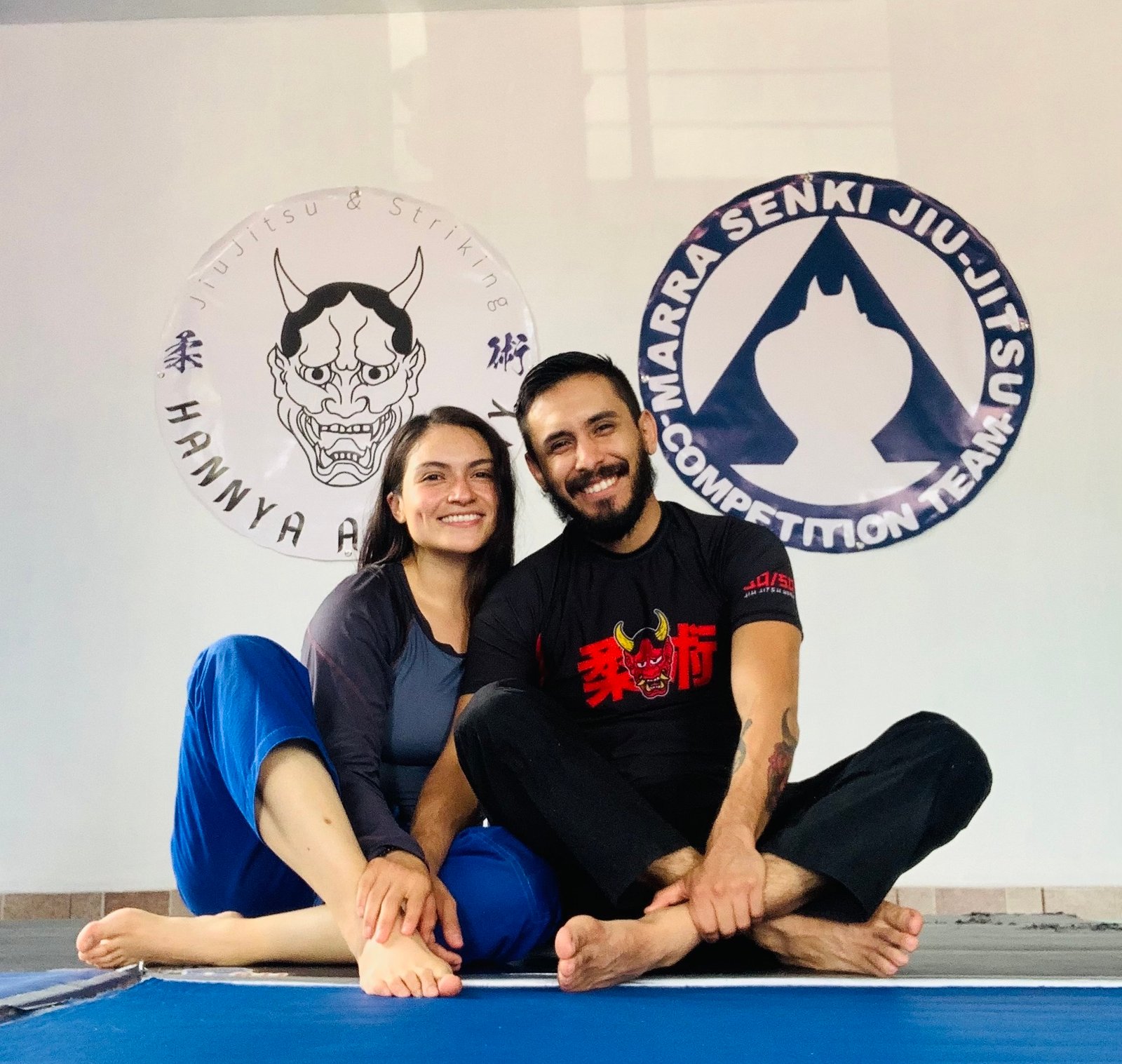Una pareja de practicantes de jiujitsu posando orgullosos sobre sus tatamis y en el fondo se ven sus logos de academia y equipo
