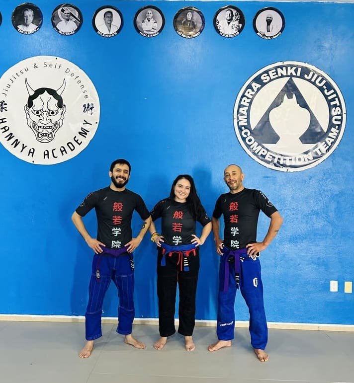 3 Profesores de jiujitsu brasileño posando para la cámara, una profesora al centro y dos profesores en cada lado, al fondo sus logos de academia y equipo de jiujitsu brasileño