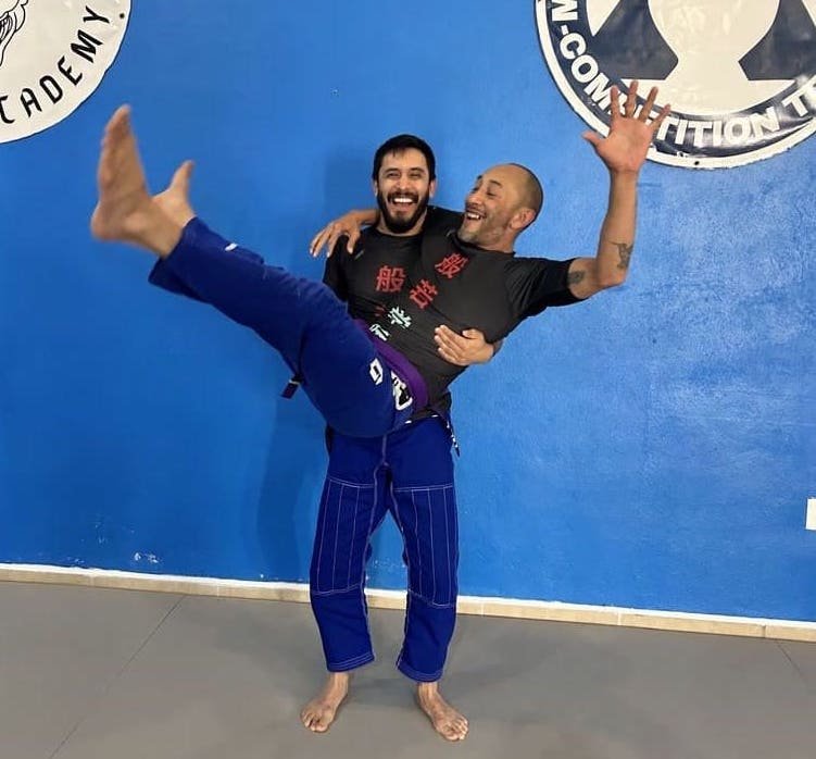 Dos profesores de jiujitsu, uno cargando al otro, ambos sonrientes en un fondo azul